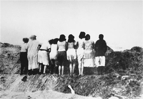 A group of Jewish women stand at the edge of an open mass grave facing the sea